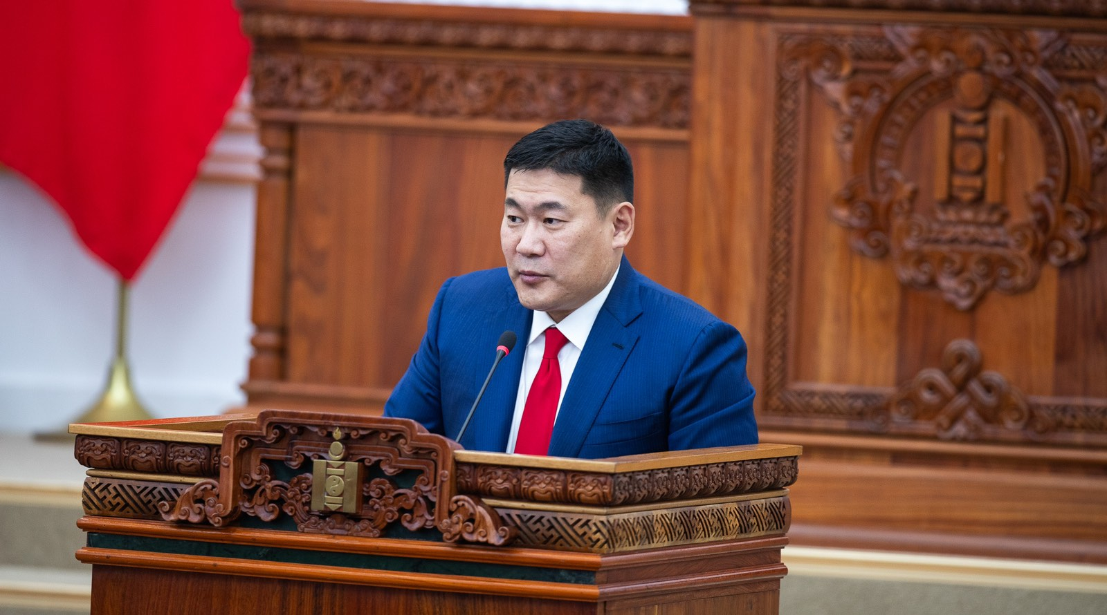 A person in a blue suit and red tie stands at a wooden podium with intricate carvings, speaking into a microphone. A red flag is partially visible in the background. The setting appears formal, possibly a government building.