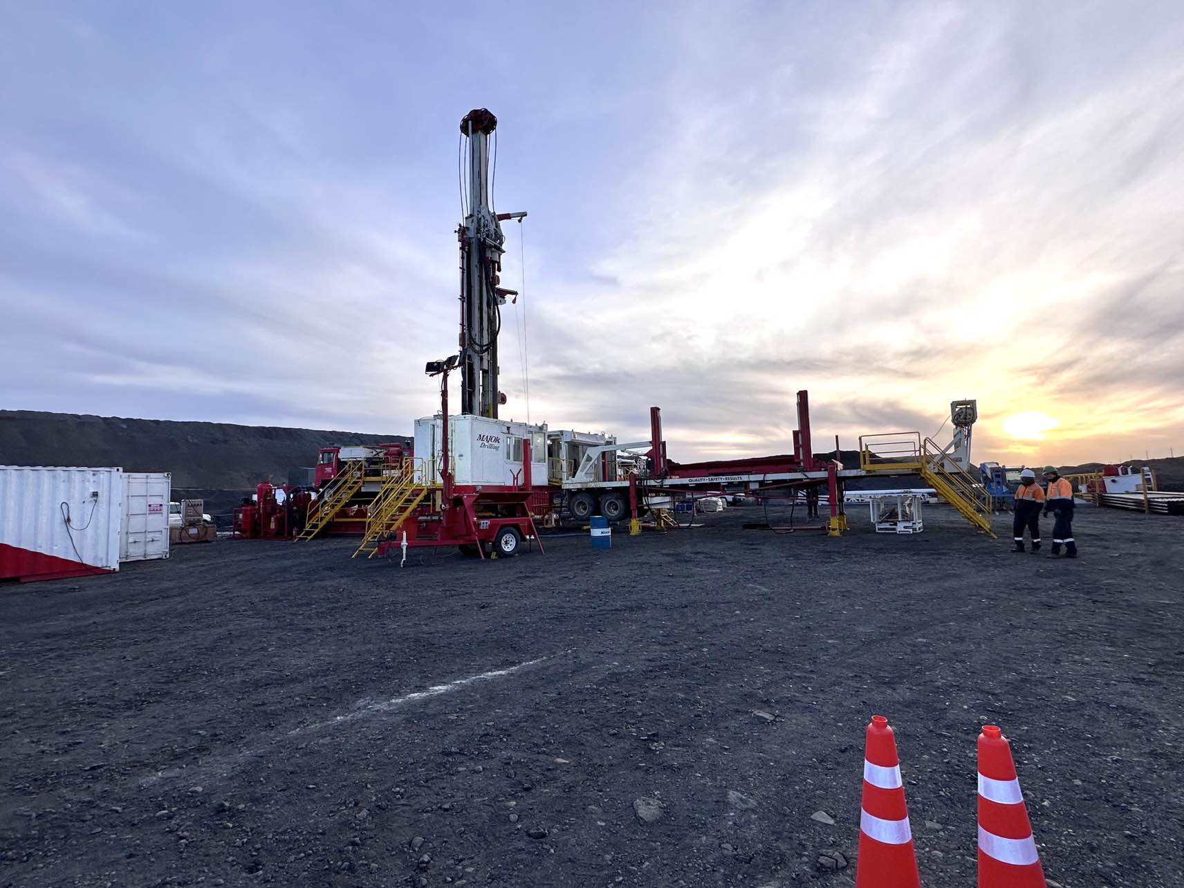 A large drilling rig stands on a gravel site with a cloudy sky and setting sun in the background, heralding эрчим хүчний салбарт гэрэл асаах мэдээ. Workers in safety gear gather around equipment and vehicles, while orange traffic cones are visible in the foreground.