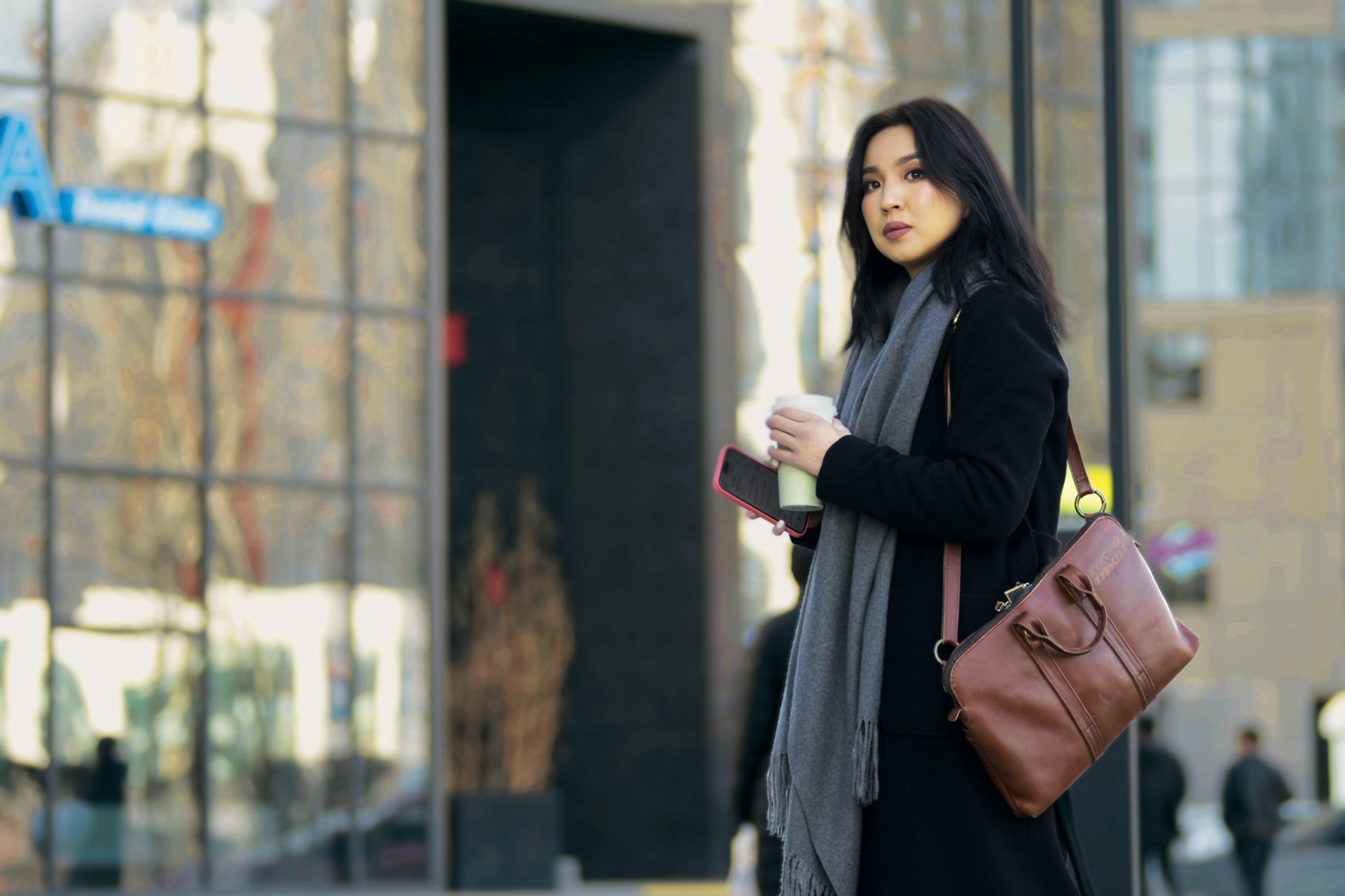 A person with long dark hair stands outside an office building, holding a coffee cup and phone. They wear a black coat, gray scarf, and carry a brown bag filled with Экспортын эрдэнэс. Reflections of trees and buildings shimmer in the glass behind them.