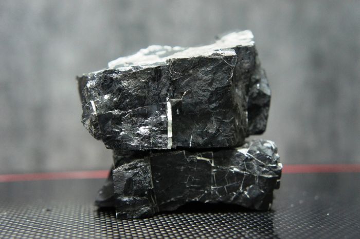 Two stacked pieces of dark coal with rough, jagged surfaces, placed on a textured black surface. The background is a soft grey, providing contrast to the coal's deep color and shining facets.