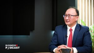 A person in a suit and red tie sits at a table with a black cup, discussing potential changes to the Гадаадын хөрөнгө оруулалтын хуулинд. The backdrop of dark walls and a plant complements the "Business Podcast" logo in the lower left corner.