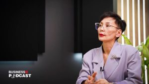 A person with short hair and glasses is seated at a table, wearing a lavender blazer with a brooch. They are speaking or listening intently, possibly during a podcast recording. A plant and a business podcast logo are visible in the background.