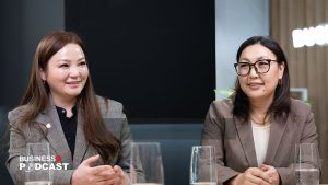 Two women in business attire sit at a table during a podcast recording. Both are smiling; one has long hair and the other wears glasses. Microphones and glasses of water are visible on the table. The setting is a modern office studio.