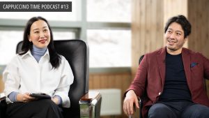 Two people sitting in armchairs engaged in conversation, one wearing a white blouse and the other a maroon jacket. A sign in the corner reads "Cappuccino Time Podcast #13.