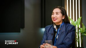 A person in a blue blazer and scarf sits at a table, smiling, with a podcast logo visible in the corner. The background is dimly lit with a plant and striped wall feature.