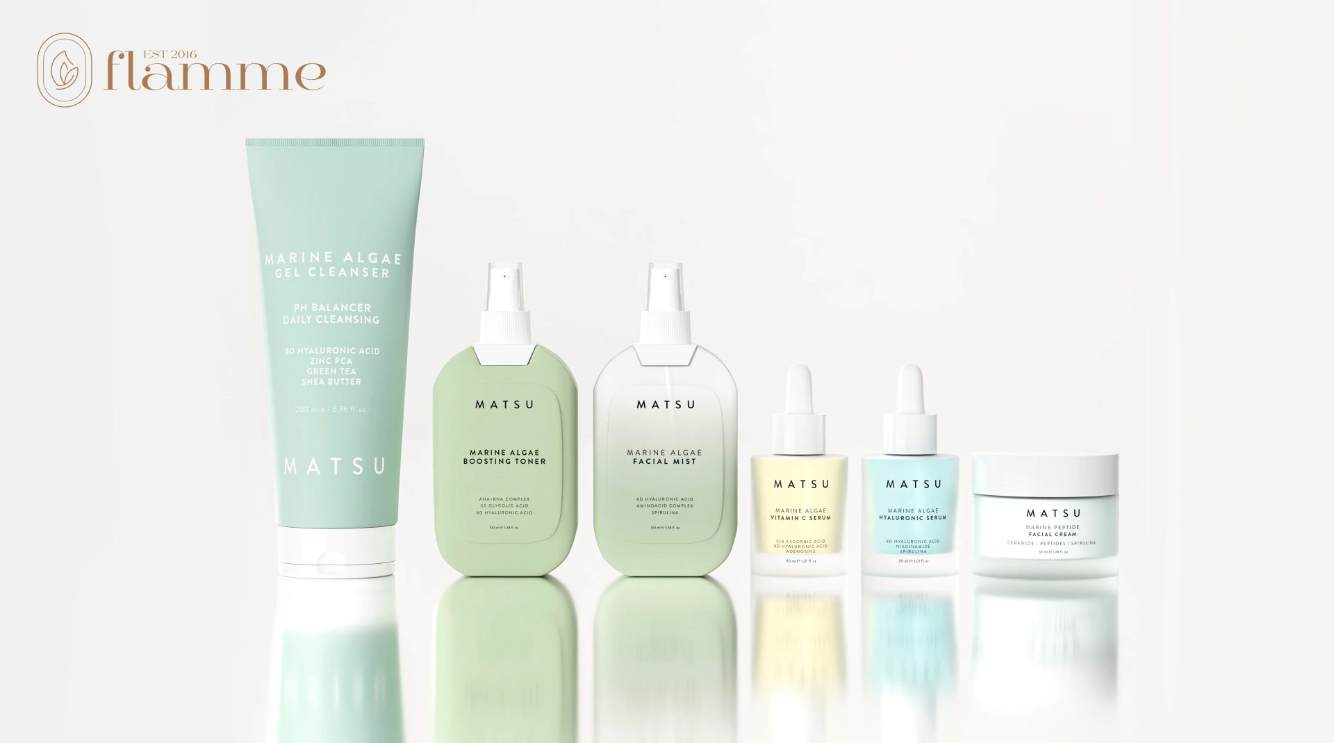 A collection of Matsu skincare products, including a gel cleanser, boosting toner, facial mist, and various serums, displayed on a white surface. The packaging is in pastel shades of green, blue, and clear bottles. Flamme logo visible in the corner.