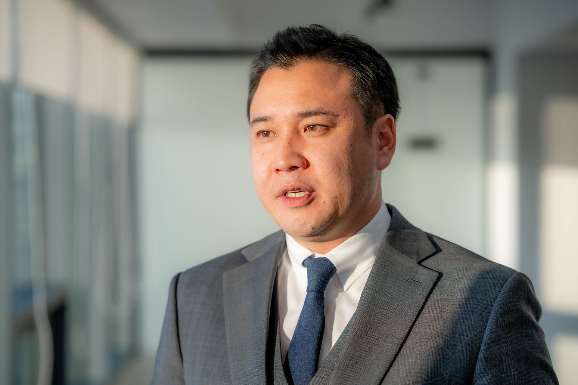 A man in a gray suit and blue tie stands in a well-lit hallway. His expression is focused and the sunlight partially illuminates his face. The background is blurred, featuring glass walls and natural light.