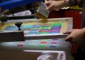 A person is using a screen printing machine to print colorful designs on a white fabric. The design includes multiple colored letter "S" shapes in a grid pattern. The person's hands are adjusting the screen.