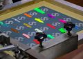 A screen printing setup with colorful ink samples applied on a flat surface. The samples include various shades like orange, pink, green, and blue, all placed on a background patterned with a circular logo. Equipment parts are visible in the foreground.