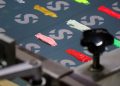 Close-up of a screen printing setup with colorful ink blobs in pink, yellow, white, green, and red on a fabric marked with repeated gray "S" symbols. A mechanical component is visible in the corner.