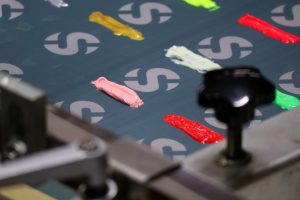 Close-up of a screen printing setup with colorful ink blobs in pink, yellow, white, green, and red on a fabric marked with repeated gray "S" symbols. A mechanical component is visible in the corner.