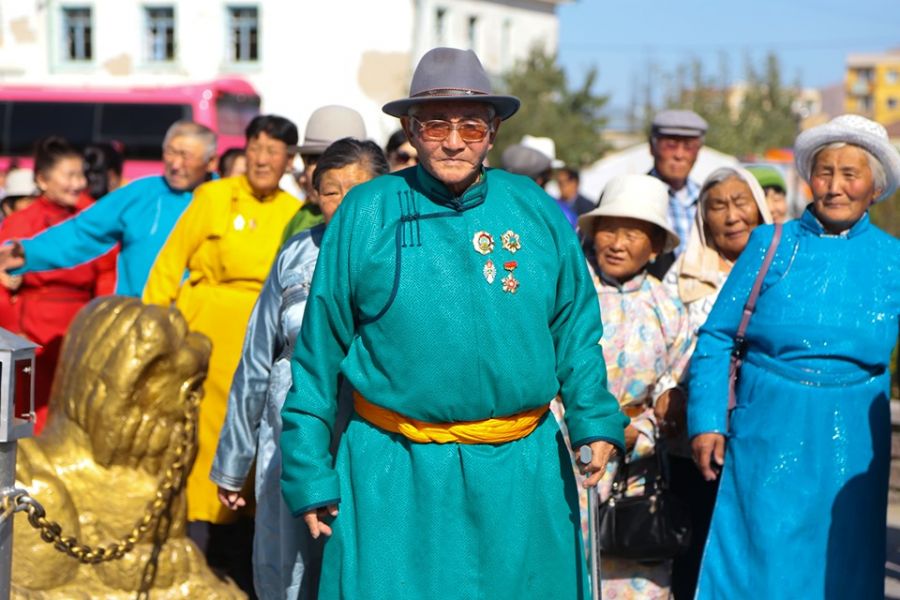 A group of elderly people wearing colorful traditional Mongolian clothing and hats stand outdoors. One man, wearing a gray hat and teal robe, is prominent in the foreground. A golden statue is partially visible to the left.