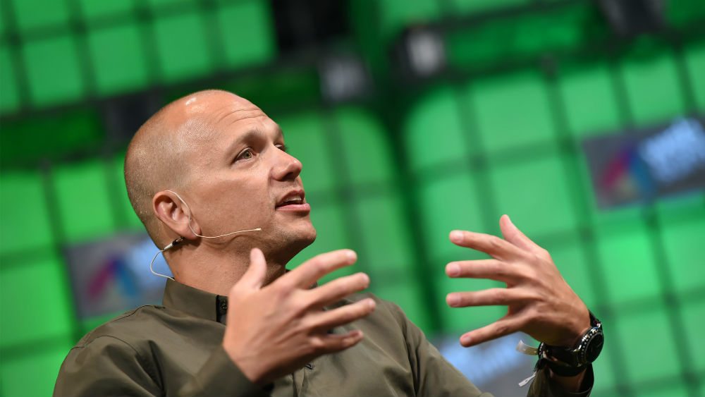 A person with a shaved head wearing a dark shirt and headset microphone is speaking on stage. Their hands are raised expressively. The background is a green grid with blurred logos.