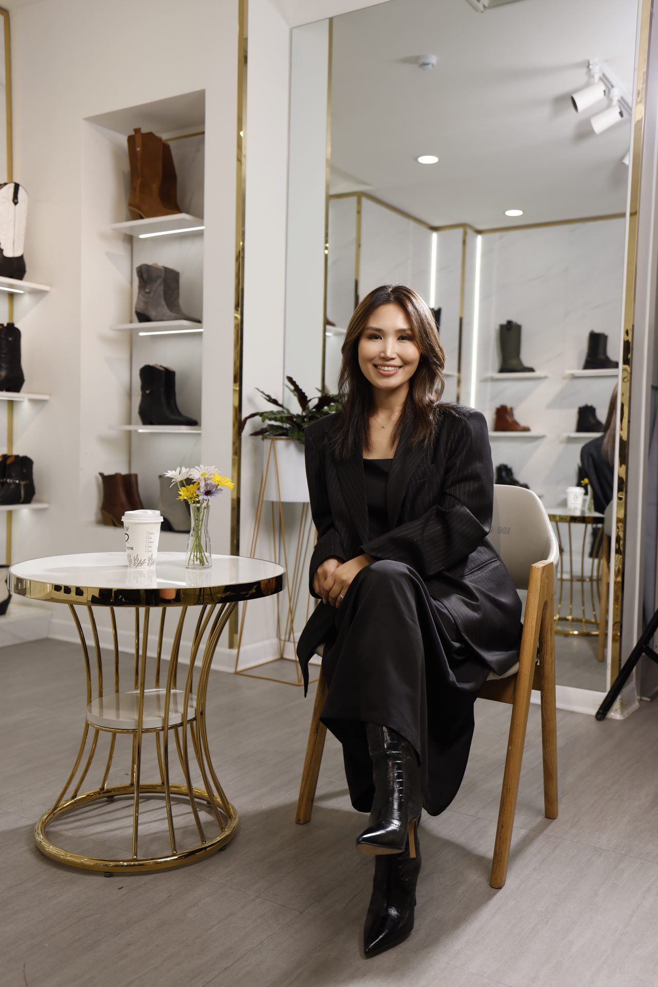 A person sitting in a modern boutique, wearing a black outfit and ankle-length boots, smiling at the camera. A round table with flowers and a coffee cup is nearby. Shelves displaying boots are visible in the background.