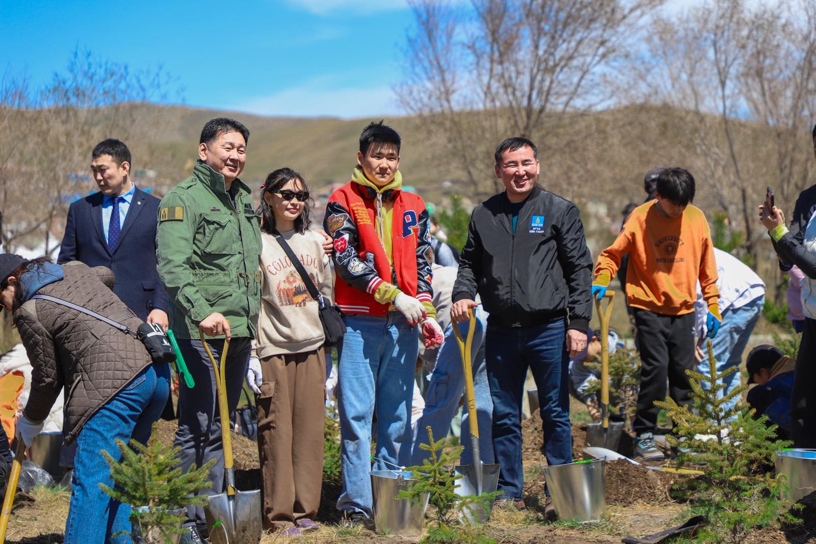 People are planting trees on a sunny day, using shovels and metal buckets. They are outdoors in a field with small trees and distant hills in the background. Some are smiling and posing for the camera.