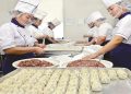 A group of chefs in uniform and hairnets are preparing dumplings in a kitchen. Two large bowls of meat mixture are placed on the table. Rows of uncooked dumplings are neatly arranged on trays. The chefs are focusing on their work.