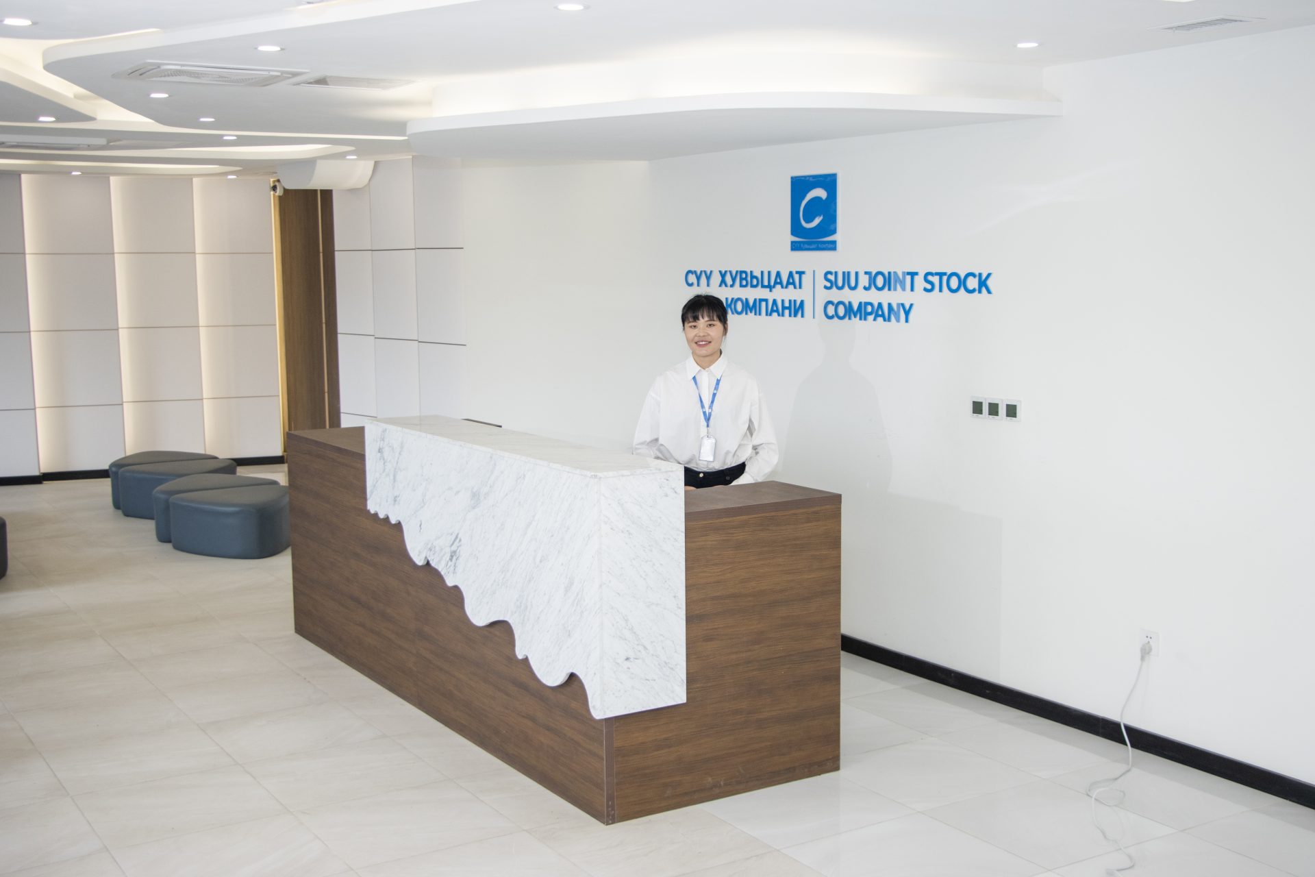 A person stands behind a reception desk in a modern office lobby. A sign above reads "SUU JOINT STOCK COMPANY" with a logo. The area is well-lit with a minimalist design and seating is visible to the left.