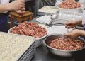 Chefs preparing dumplings in a kitchen. Bowls of minced meat are being used as filling. Several trays hold rows of assembled dumplings, and dough circles are visible in the background. Hands are working with spoons and dough.