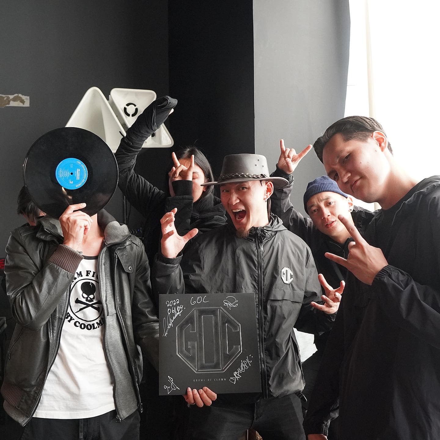 A group of five people posing playfully indoors. One person covers their face with a vinyl record, while others make rock signs and smile. They hold a signed album in the center. The background features a dark wall and some ambient light.