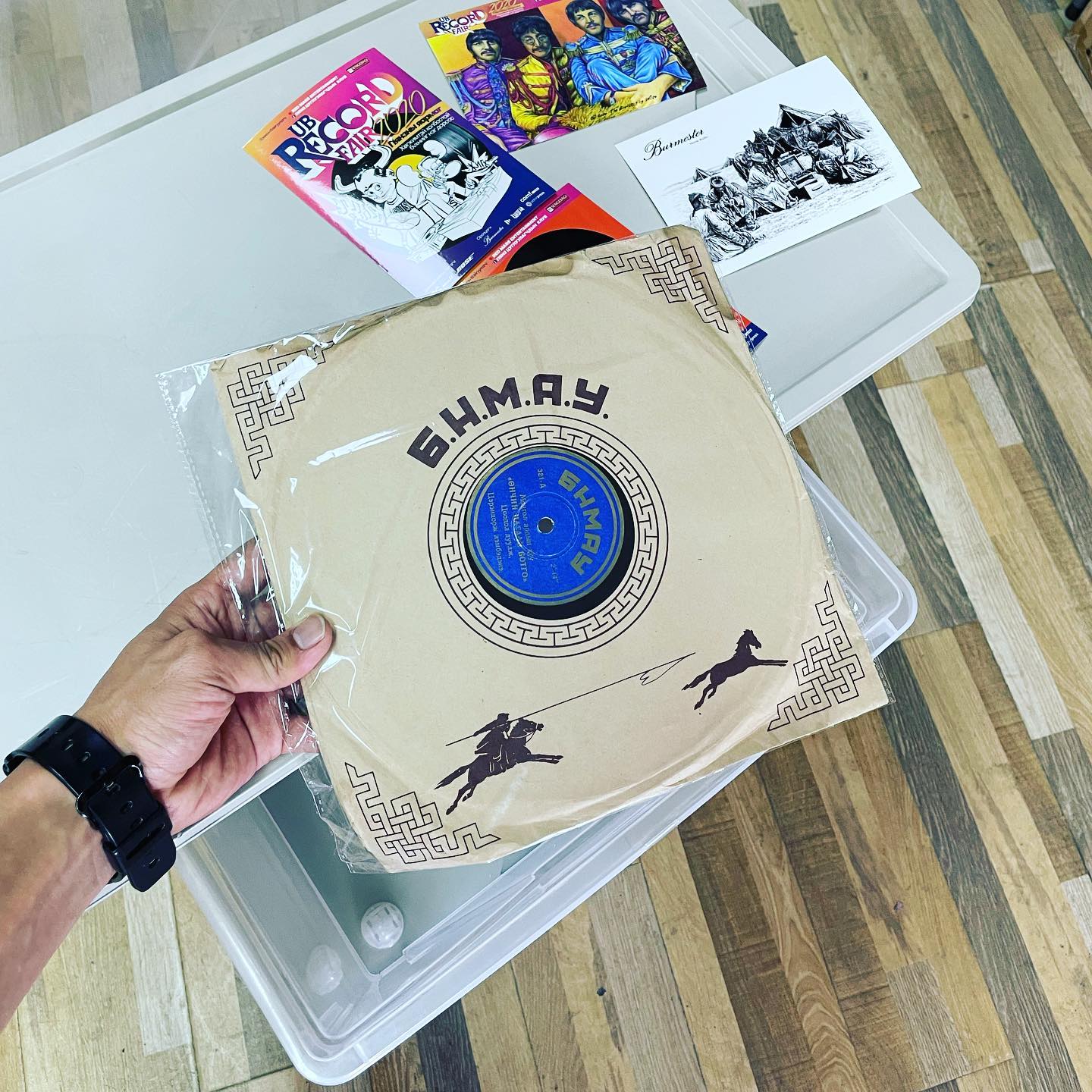 A hand holds a vintage vinyl record with Cyrillic letters on the cover, a design featuring a warrior on horseback. Surrounding the record are comic books and postcards on a wooden floor, next to a transparent plastic container.