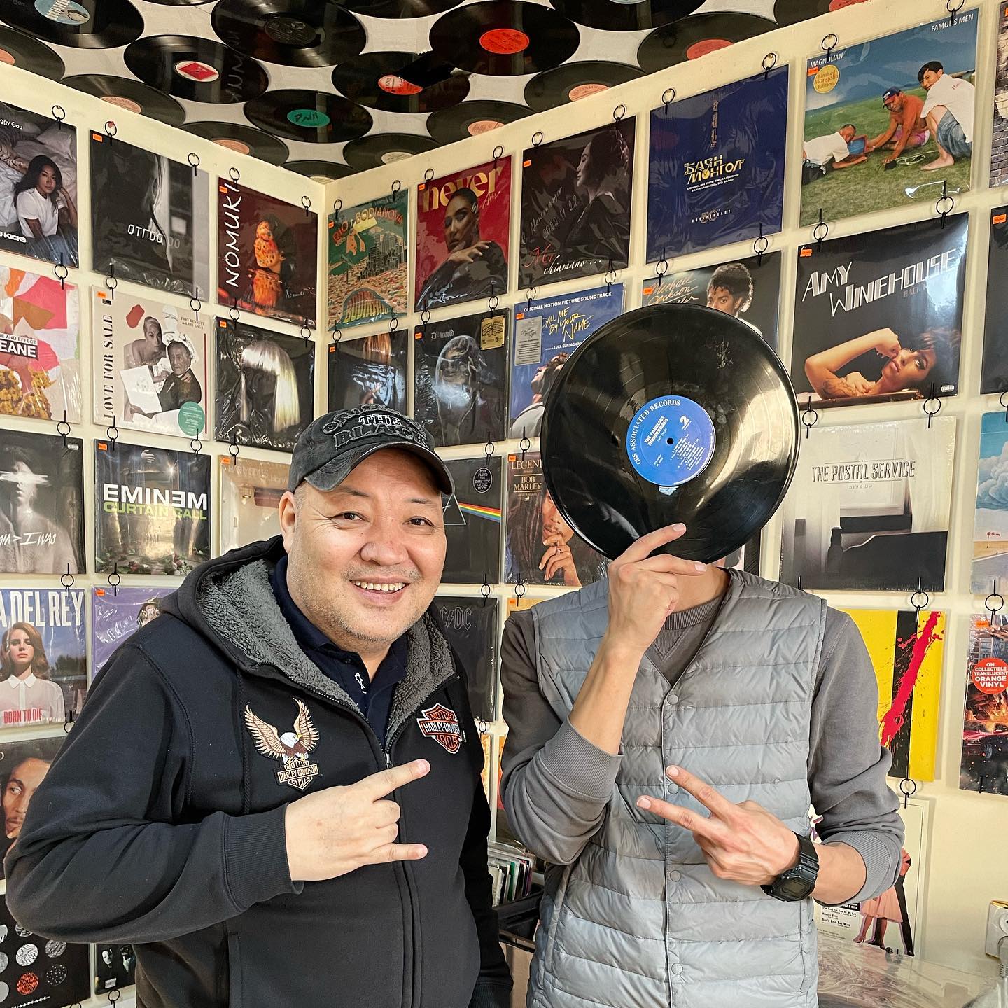 Two people standing in a room with vinyl records on the walls. The person on the left is smiling and gesturing with his hand. The person on the right holds a vinyl record in front of their face, also gesturing with his hand.