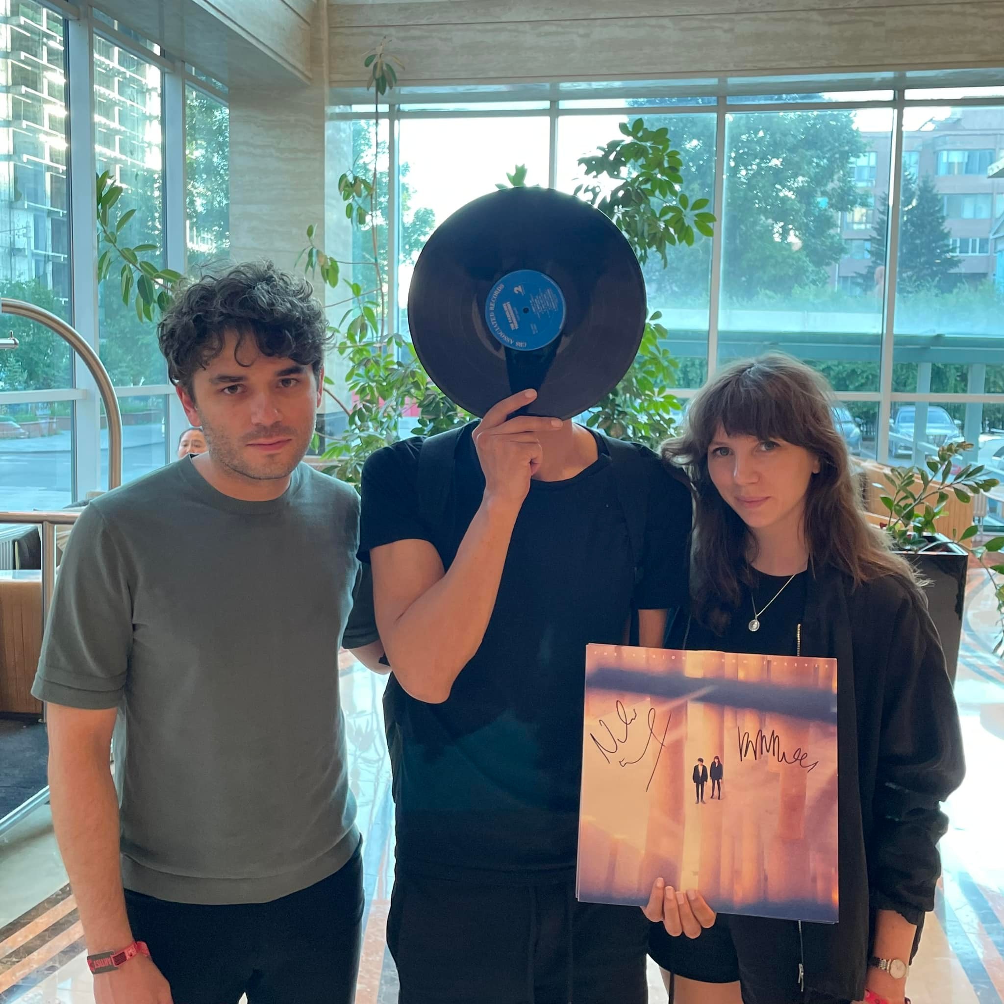 Three people standing indoors. The person in the middle holds a vinyl record over their face and an album cover signed by artists. The other two people stand on either side, smiling slightly. Green plants and large windows are in the background.