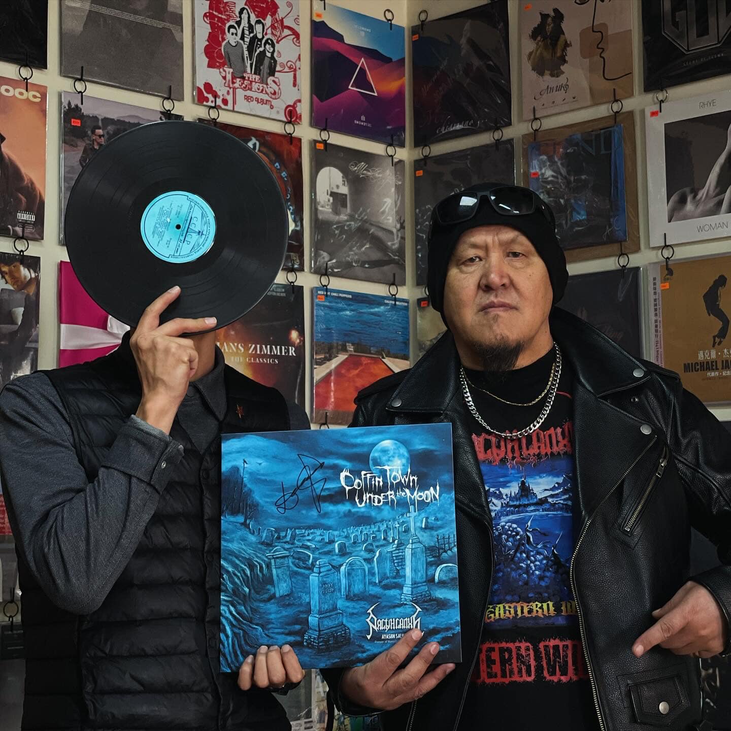 Two people pose in a music-themed room. One holds a vinyl record over their face, while the other wears a beanie and leather jacket, holding an album cover titled "Coffin Town." Various album covers are displayed on the wall behind them.