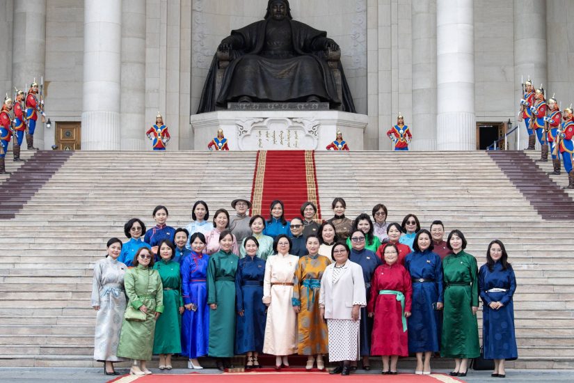 A group of women in colorful traditional outfits stand on the steps of a large building with a statue. The scene includes a red carpet and uniformed guards on both sides.