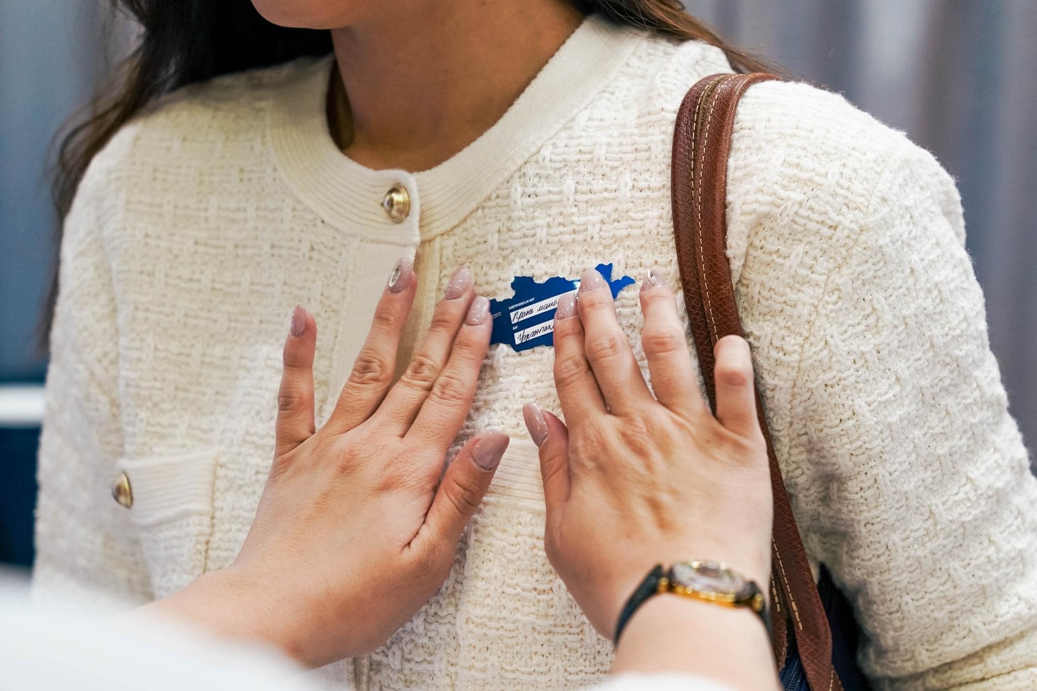 A person affixes a blue name tag onto the lapel of another person wearing a white textured jacket. The scene is close-up and focused on the hands and the clothing, with blurred background details.