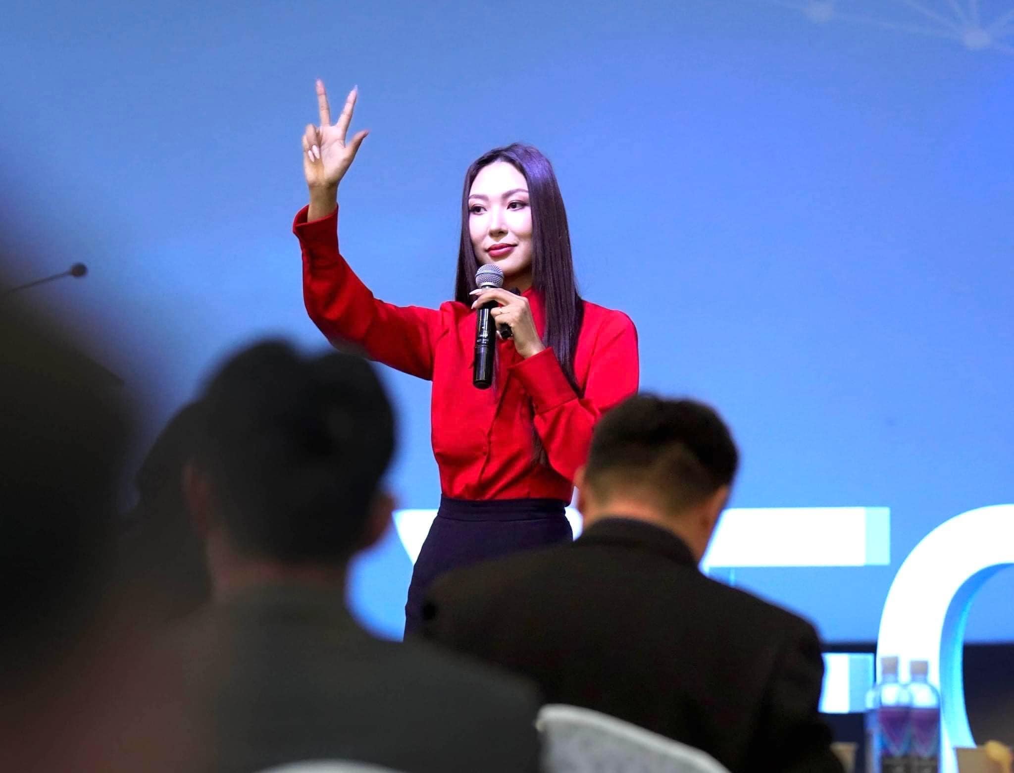 A woman in a red blouse stands on stage holding a microphone, raising three fingers. She is speaking to an audience, with a blurred view of attendees in the foreground. The background is a blue screen.