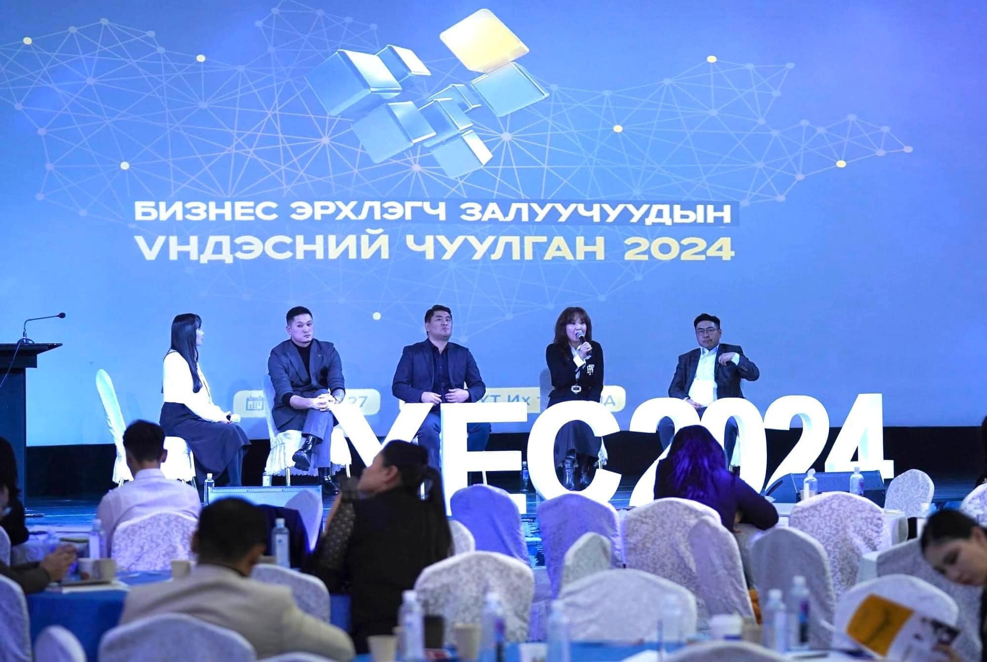 Five people sit on a stage, engaged in a panel discussion, with "Business Entrepreneur Youth National Forum 2024" written behind them in a mix of languages. The audience listens attentively in a large conference room.