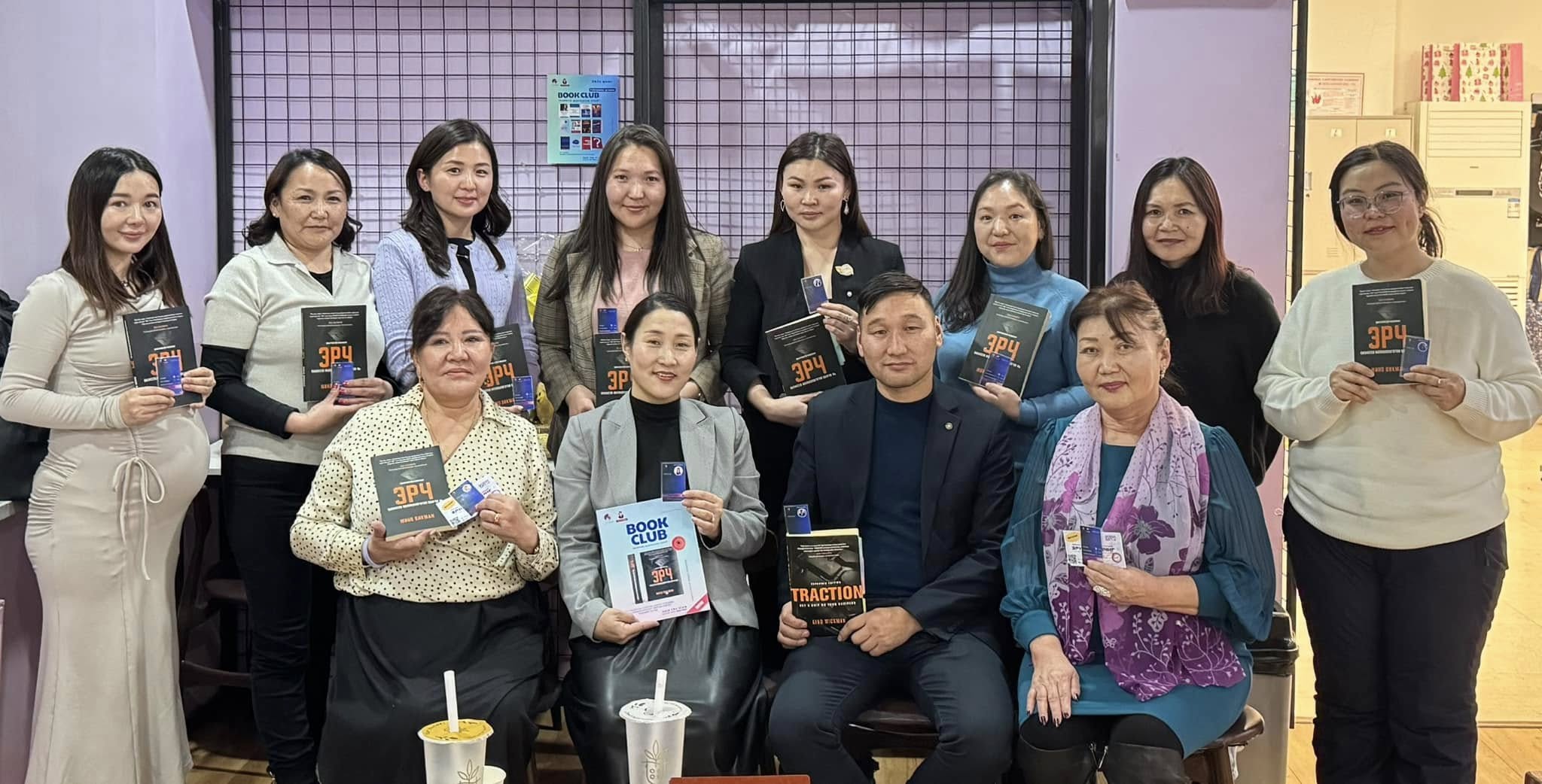 A group of people posing together in a room, each holding different books. Some are seated while others stand behind them. They are smiling, and the setting appears casual, with a couple of drinks visible on a table in front.