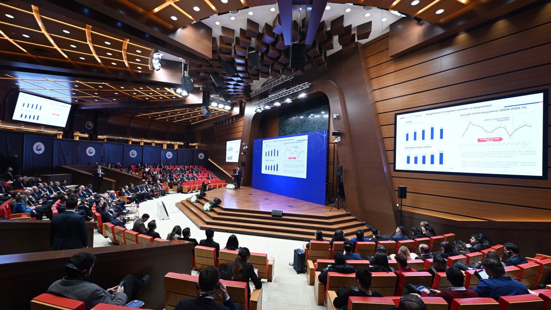 A modern auditorium with wooden paneling and red seating is filled with people attending a presentation. Large screens display graphs and charts, while a speaker is at the front. The high ceiling features unique geometric patterns and recessed lighting.