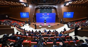 A large conference room filled with attendees seated in red chairs. A speaker stands at a podium on a stage with a blue backdrop displaying two flags. Several cameras and media personnel are visible in the foreground.