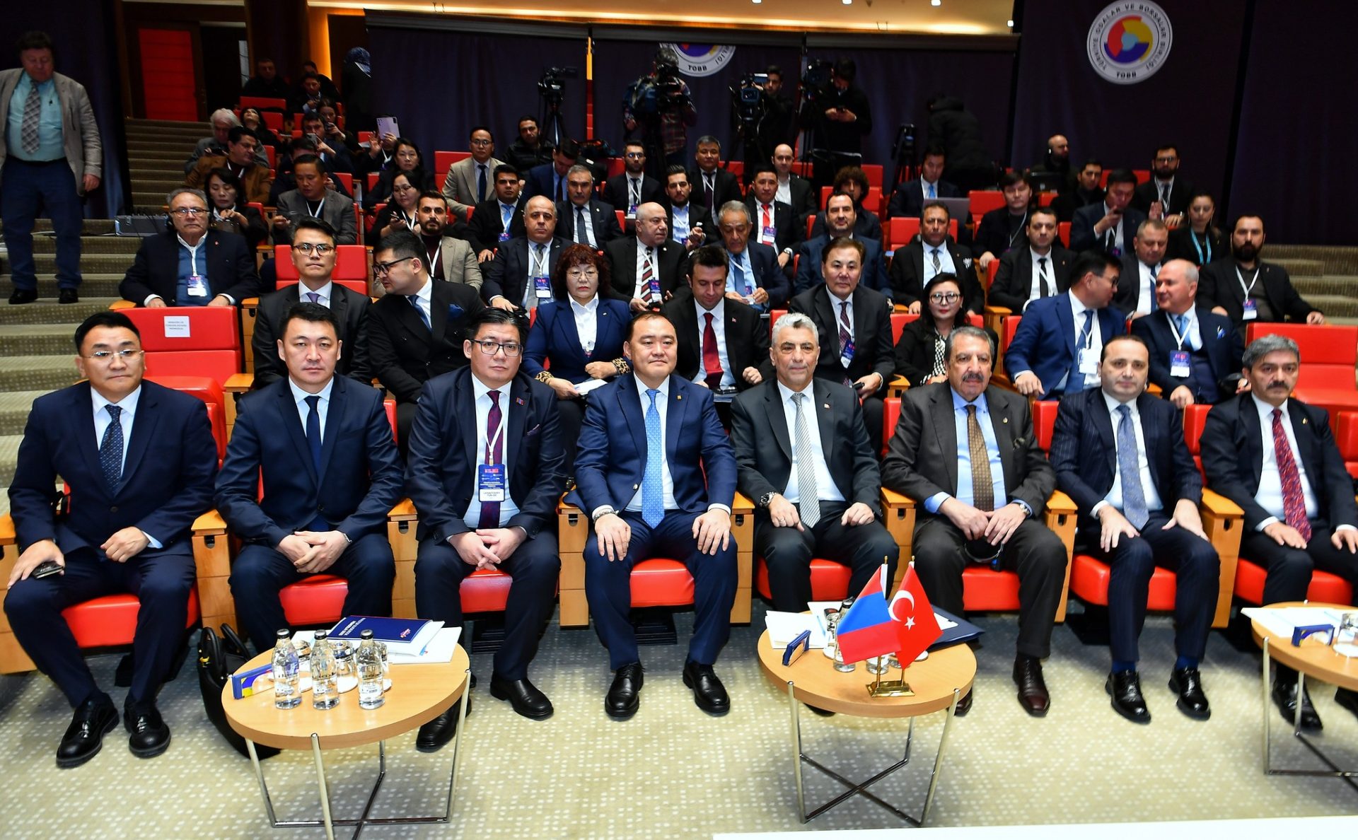 A group of men in suits sit in rows of red chairs, facing forward. A Turkish flag is on a table at the front. People are seated behind, with some standing. The setting appears to be a formal conference or event hall.