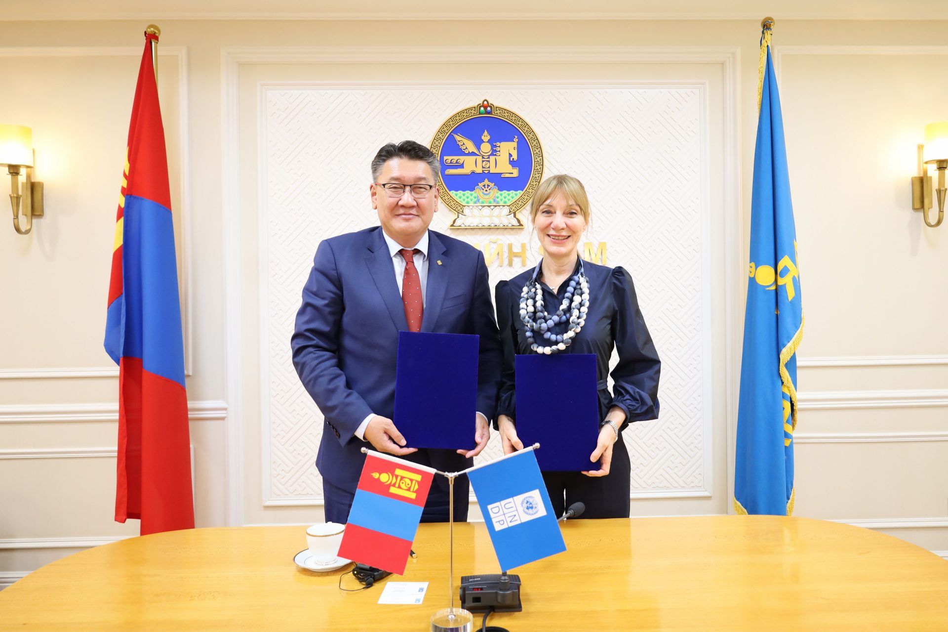 Two individuals stand side by side, holding documents, in a formal setting. Flags, including Mongolia's, are in the background. A table with a small Mongolian flag and a coffee cup is in the foreground.