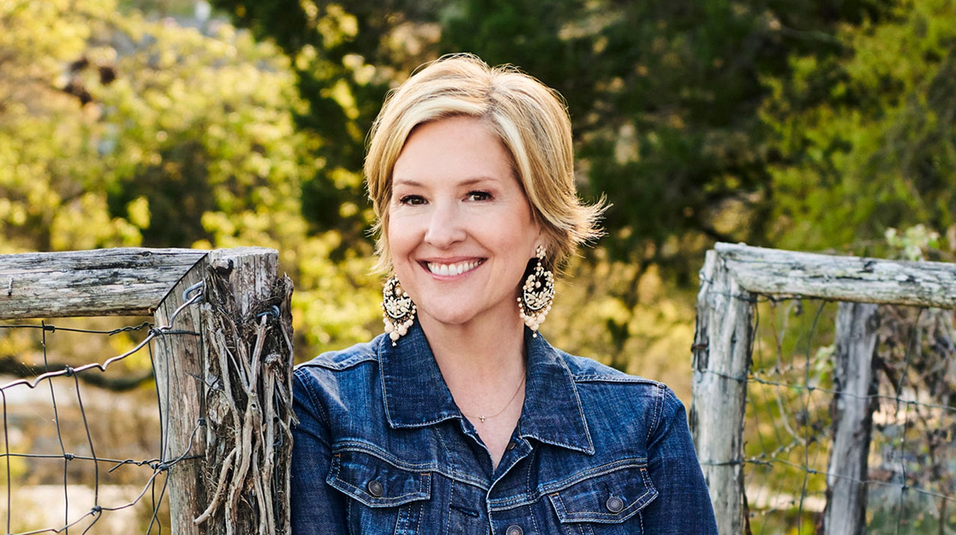 A person with short blonde hair wears a denim jacket and large earrings, smiling outdoors. They are leaning on a rustic wooden fence with a backdrop of green trees.