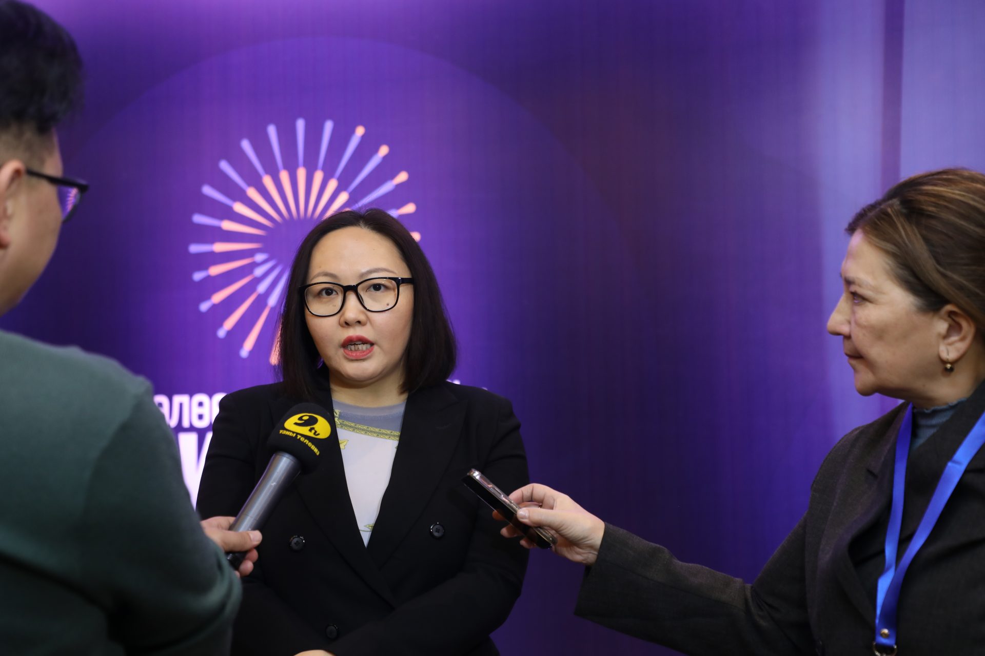 A woman in a black blazer speaks in front of microphones held by two reporters. She stands against a purple backdrop with a circular, radiating pattern. One reporter is holding a yellow microphone.