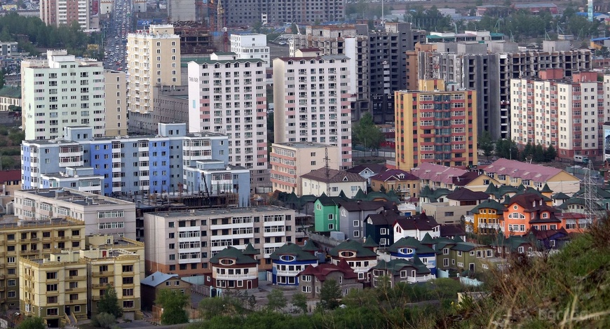 A cityscape showcasing a lively mix of colorful modern and older multistory buildings and houses, echoing the Өнгөрсөн сарын өсөлт ба бууралт. Vibrant houses dominate the foreground, while taller apartment buildings rise behind them, with green trees interspersed throughout.