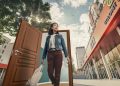 A woman carrying a bag steps through an open door into an outdoor urban scene, epitomizing the spirit of positive change. She wears a denim jacket, looking ahead with a smile. Among modern buildings and trees beneath a clear blue sky with some clouds, the prospect of ипотекийн зээл awaits.