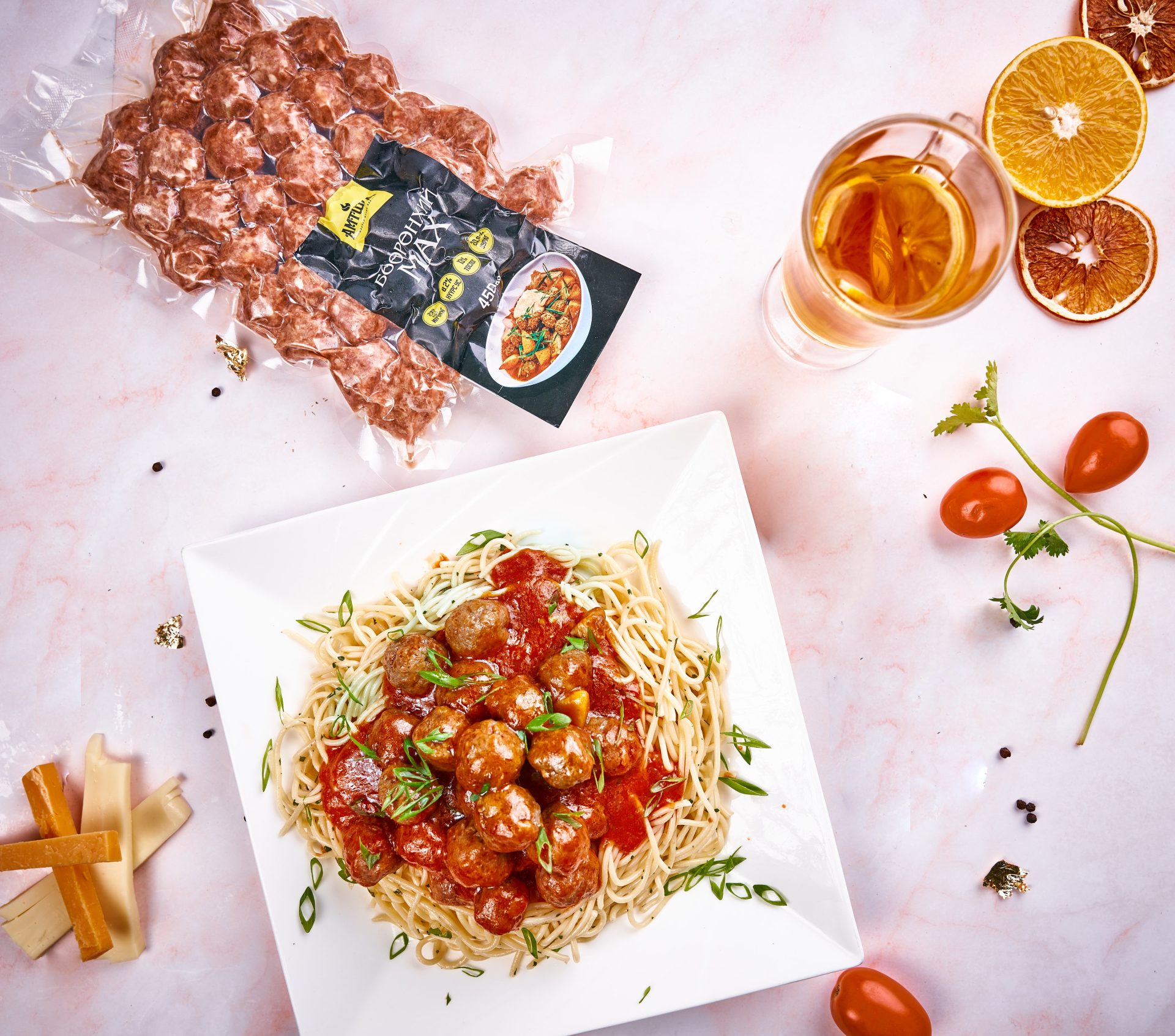 A square white plate with spaghetti and meatballs in tomato sauce garnished with herbs. Nearby are cherry tomatoes, dried orange slices, a glass of juice, and a pack of dried pasta on a pink marble surface.