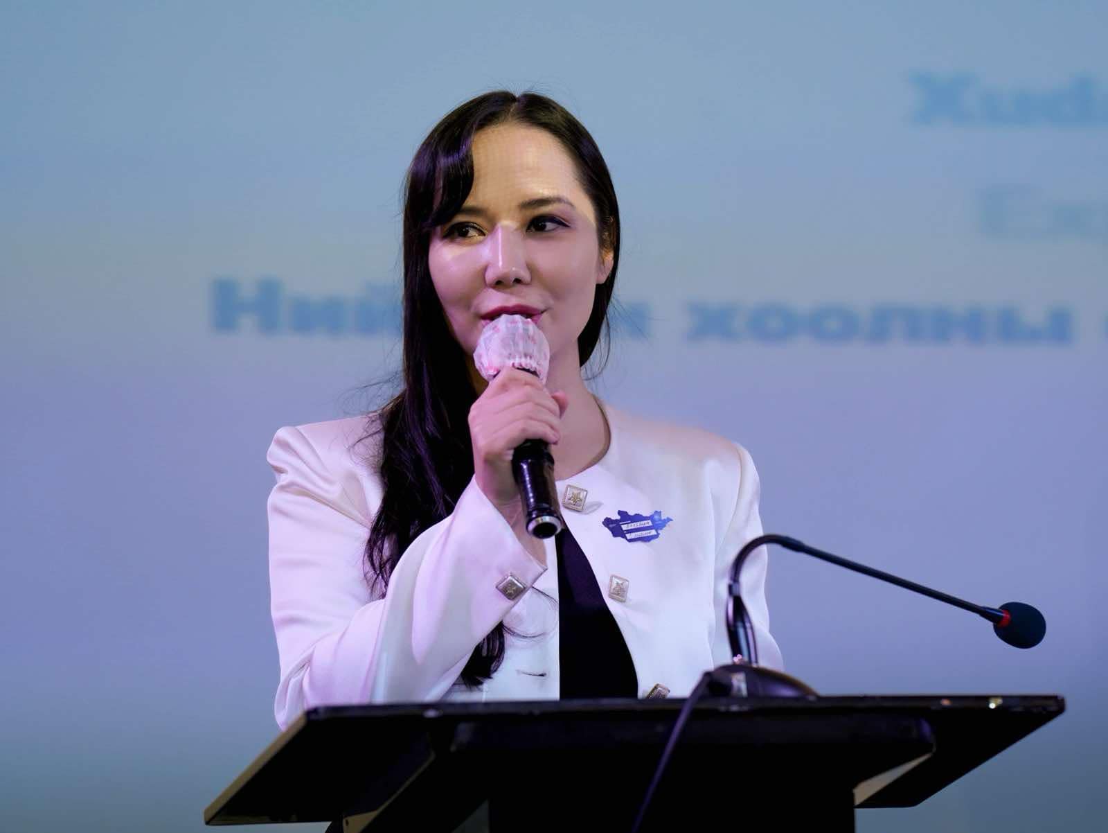 A woman in a white blazer speaks into a microphone at a podium, with a light blue screen displaying words in a non-Latin script in the background.