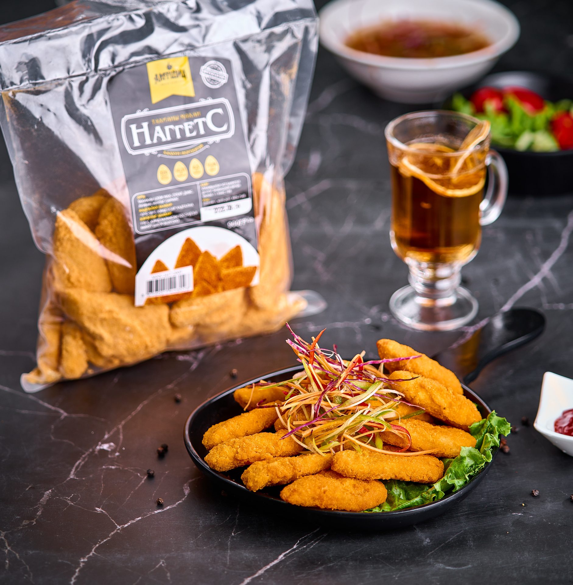 A plate of golden brown chicken nuggets garnished with sprouts sits on a marble countertop. A packet of frozen nuggets is in the background, alongside a glass of tea and a small dish of salad.