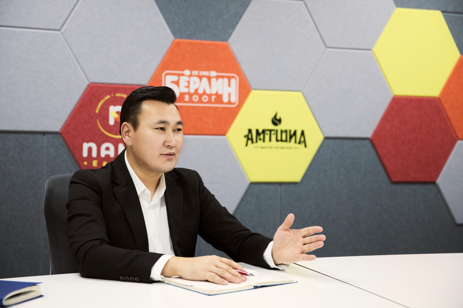 A person in a black suit sits at a table with a notebook in front of them, gesturing with one hand. The background features hexagonal wall panels in various colors with logos and text.