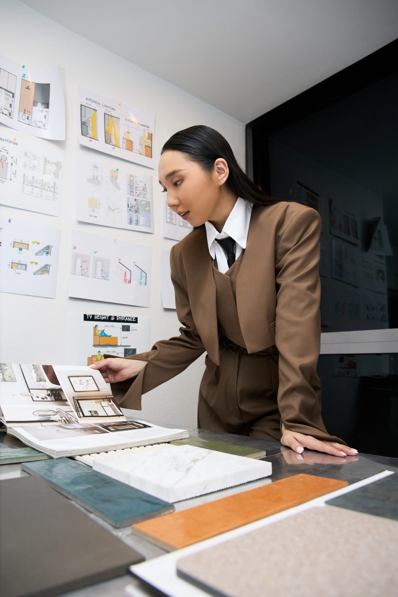 A person in a brown suit examines interior design materials and plans on a table. Various design sketches are displayed on the wall behind them.