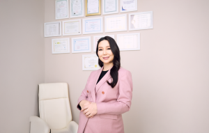 A woman with long dark hair stands confidently in a pink blazer, smiling. She is in a professional office with a cream chair and numerous framed certificates on the wall behind her.