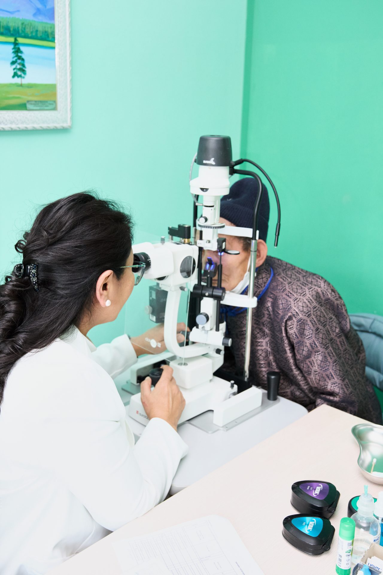 A healthcare professional conducts an eye exam on a person using a slit lamp in a well-lit room. The person being examined is seated, while the professional adjusts the equipment. Various ophthalmic tools are visible on the table.