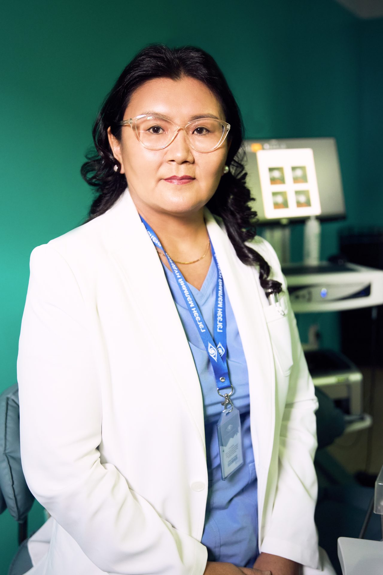 A woman in a white lab coat and glasses stands in a medical office, wearing a blue ID lanyard over her scrubs. Behind her is medical equipment. She looks directly at the camera.