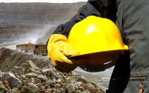 A person in safety gloves holds a yellow hard hat at a mining site. Large dump trucks are visible in the background, and layers of earth and rock formations are spread across the landscape. Dust rises in the air under a clear sky.