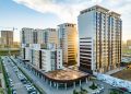 A modern urban residential complex with multiple high-rise buildings under a blue sky offers attractive ипотекийн зээл options. The sleek design features glass and beige facades, while cars are parked in neat rows along the street, as sunlight peeks through the structures.